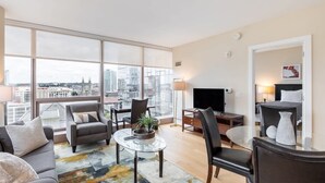 A modern living room with a sofa, chairs, a coffee table, and a TV. Large windows offer a city view.