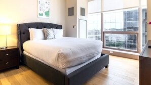 A modern bedroom with a large bed, a nightstand, a window with a city view, and a wall-mounted air vent.