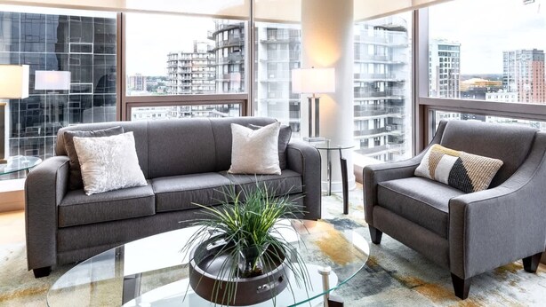 A modern living room with a grey sofa and armchair, a glass coffee table, and a view of high-rise buildings.