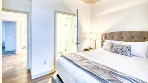 Une chambre moderne avec un grand lit, une tête de lit capitonnée, une lampe de chevet et une vue sur la salle de bain par une porte ouverte.