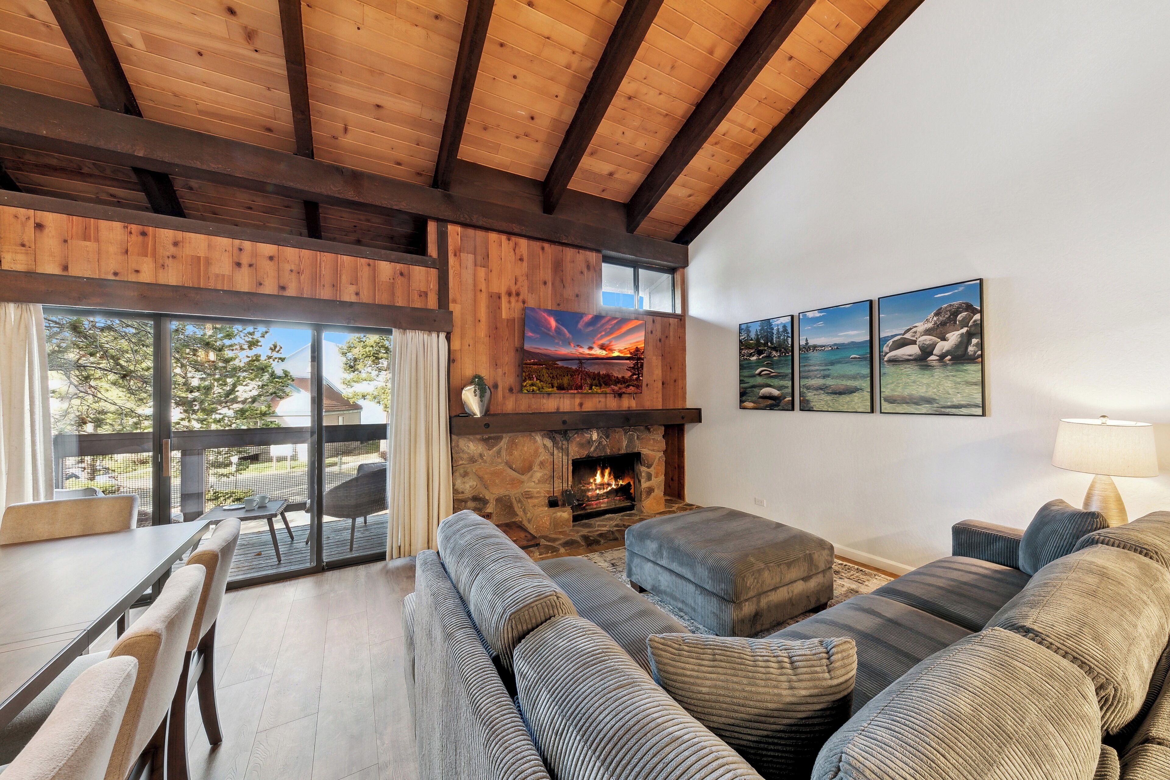 A cozy living room with a stone fireplace, wooden beams, and a large window overlooking a deck.