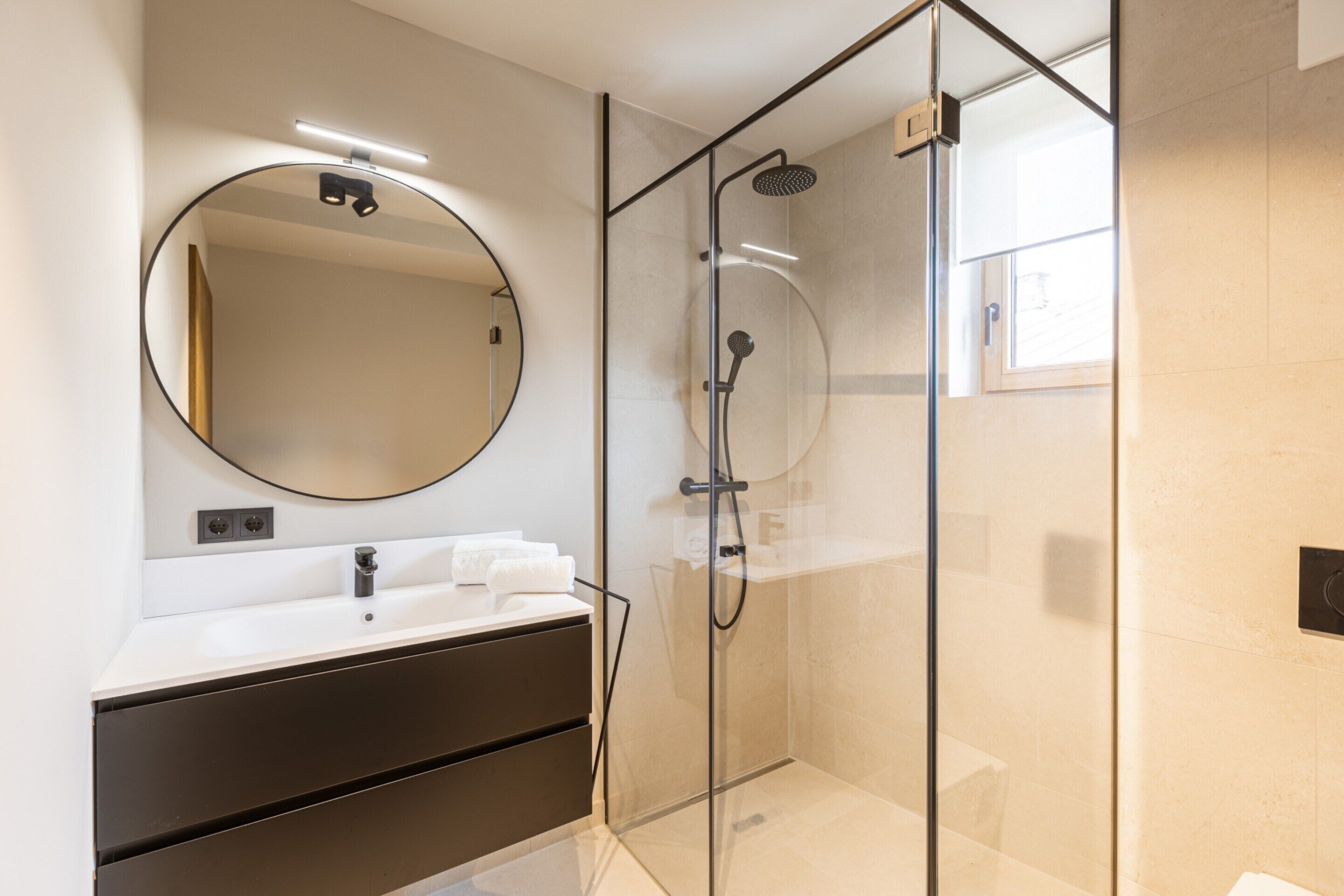 A modern bathroom with a glass-enclosed shower, a round mirror, and a white vanity with a dark cabinet.
