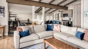 A modern living room with a sectional sofa, wooden beams on the ceiling, and a kitchen in the background.