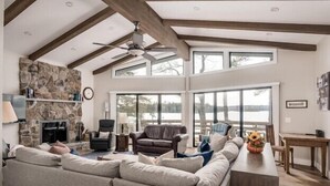 A spacious living room with a stone fireplace, large windows, and a high ceiling with exposed beams.