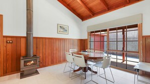 A dining area with a wood-burning stove, white chairs, and a large window with a view. - Time and Tide in Inverloch (Inverloch)