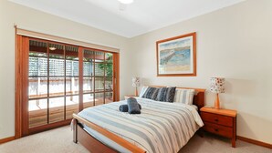 A bedroom with a large sliding glass door to a patio, a bed with striped bedding, two bedside tables with lamps, and a framed painting on the wall. - Time and Tide in Inverloch (Inverloch)