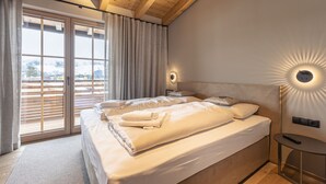 A bedroom with a large bed, a nightstand, a wall-mounted light, and a view of the outdoors through a sliding door. - Penthouse in Tyrol With Infrared Sauna (Westendorf)