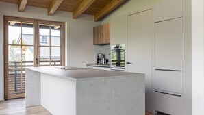 A modern kitchen with a large island, wooden ceiling beams, and a view of a house through a sliding door. - Penthouse in Tyrol With Infrared Sauna (Westendorf)