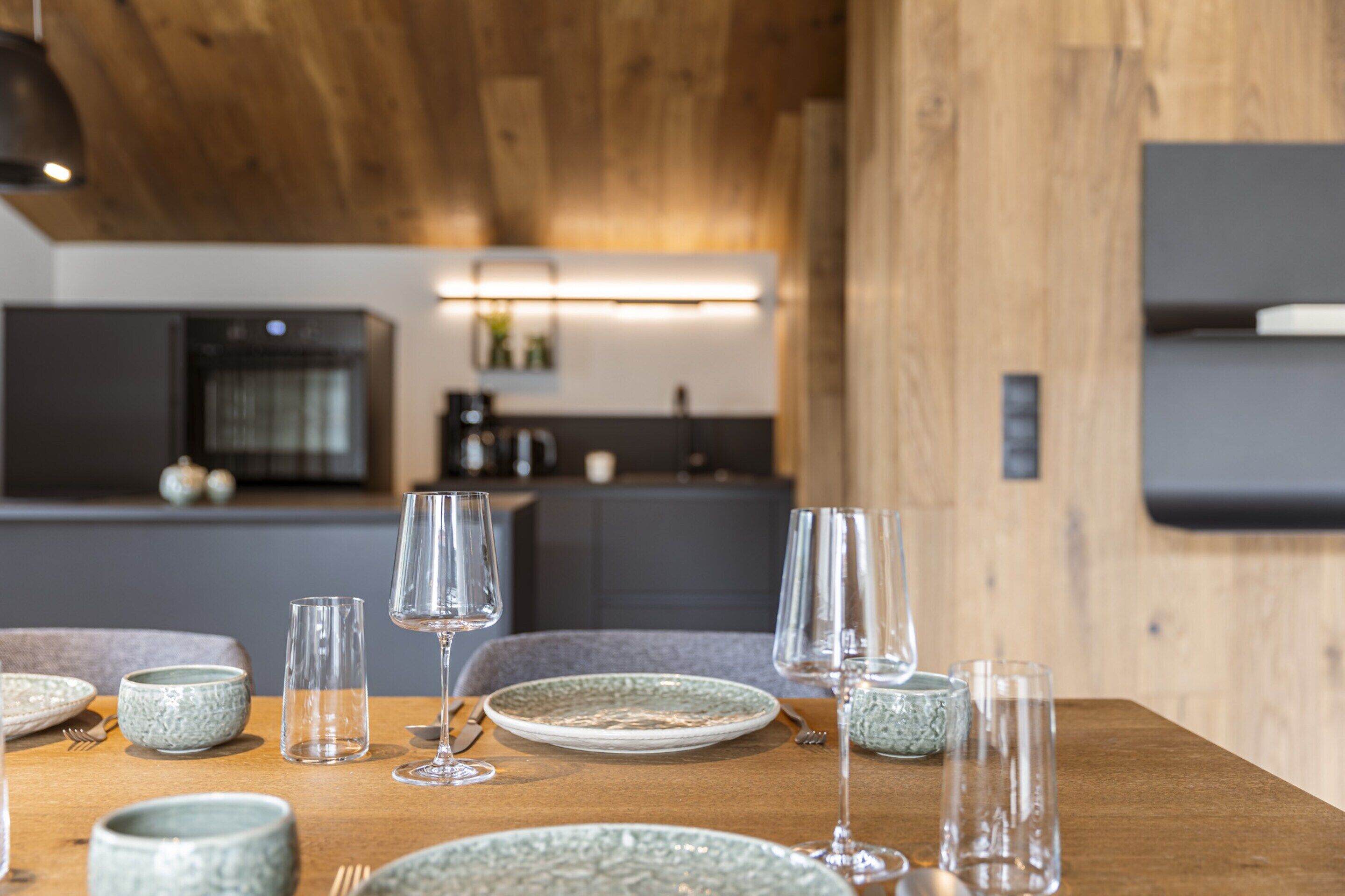 A dining table set with plates, bowls, and wine glasses, with a kitchen in the background.