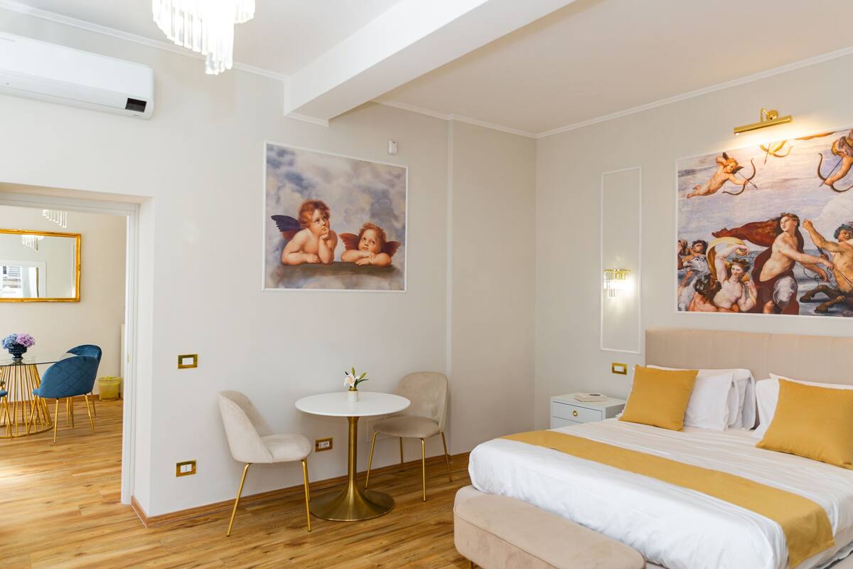 A hotel room with a bed, a round table with chairs, a desk, and a painting on the wall.