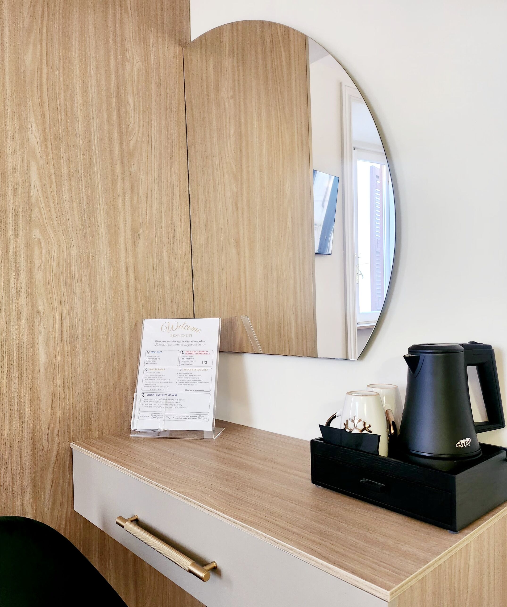 A hotel room with a wooden desk, a black tray with a coffee maker, a mirror, and a welcome sign.
