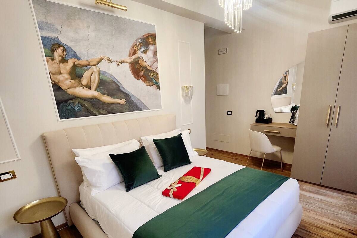 A hotel room with a large bed, a desk, a chair, a small round table, a painting on the wall, and a chandelier.