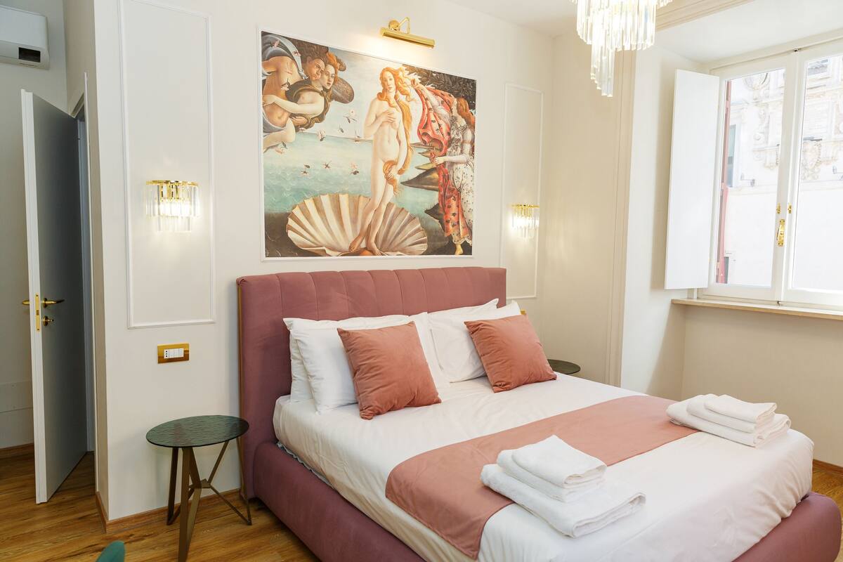A bedroom with a large bed, a small table, a painting on the wall, and a window with a view.