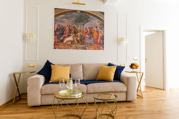 Un salon avec un canapé beige, deux tables d’appoint et une table basse en verre. Un grand tableau est accroché au mur, au-dessus du canapé.