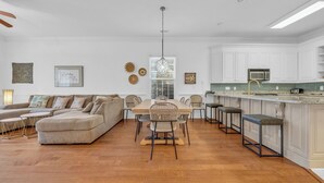 A spacious living room with a large L-shaped sofa, a dining table with chairs, and a kitchen area with white cabinets and a bar stool.