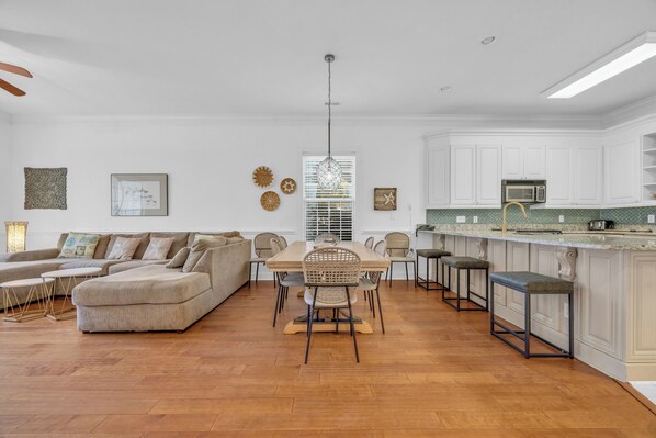 Amplia sala de estar con un sofá en forma de L, una mesa de comedor con sillas, y una cocina con armarios blancos y un taburete de bar.