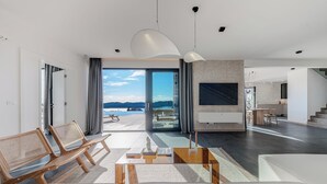 Ein modernes Wohnzimmer mit Sofa, Sesseln, Couchtisch und Fernseher. Ein großes Fenster bietet einen Blick ins Freie.