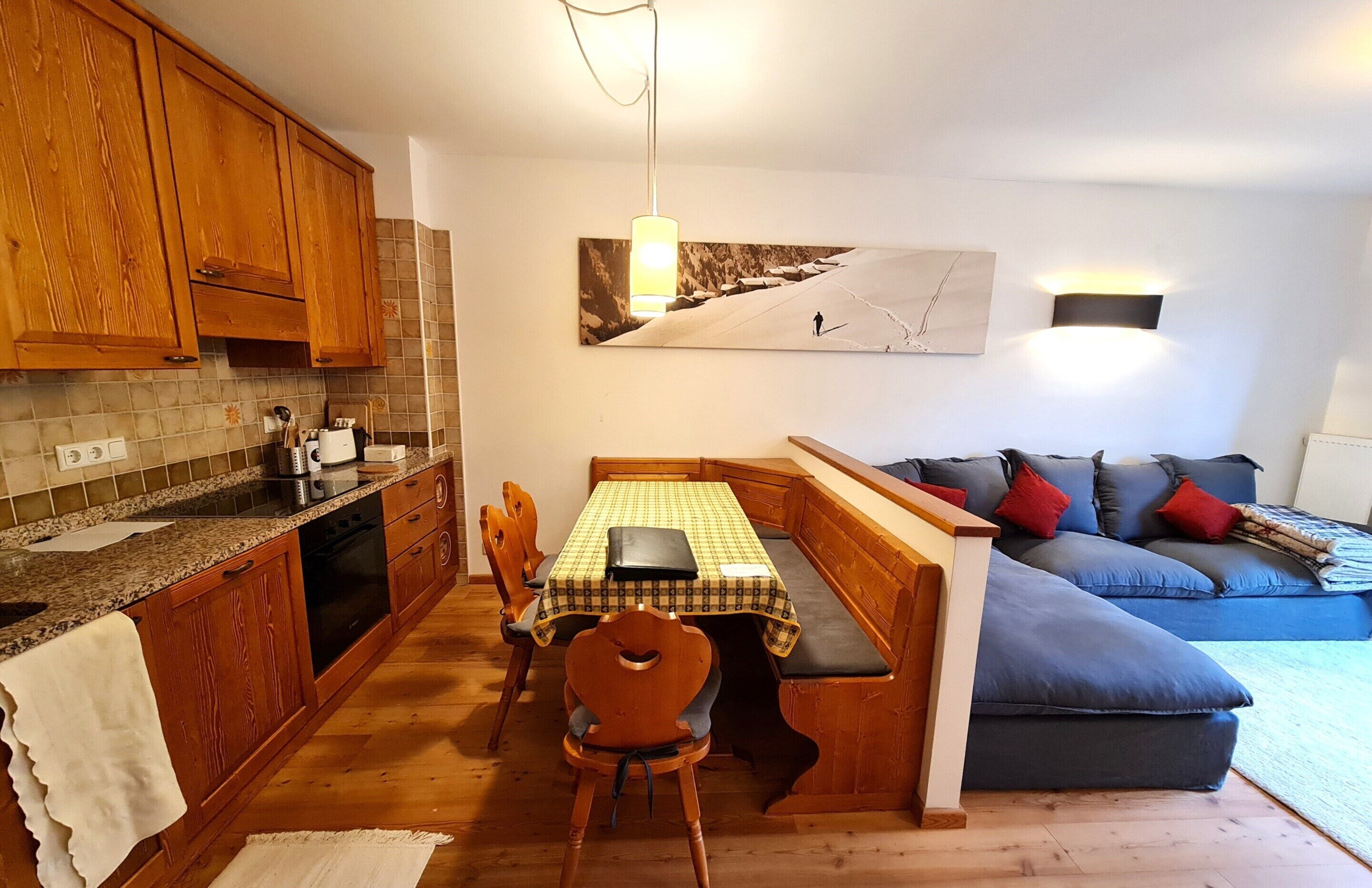 A kitchen with wooden cabinets, a dining table with chairs, a bench, a sofa, and a wall-mounted picture.