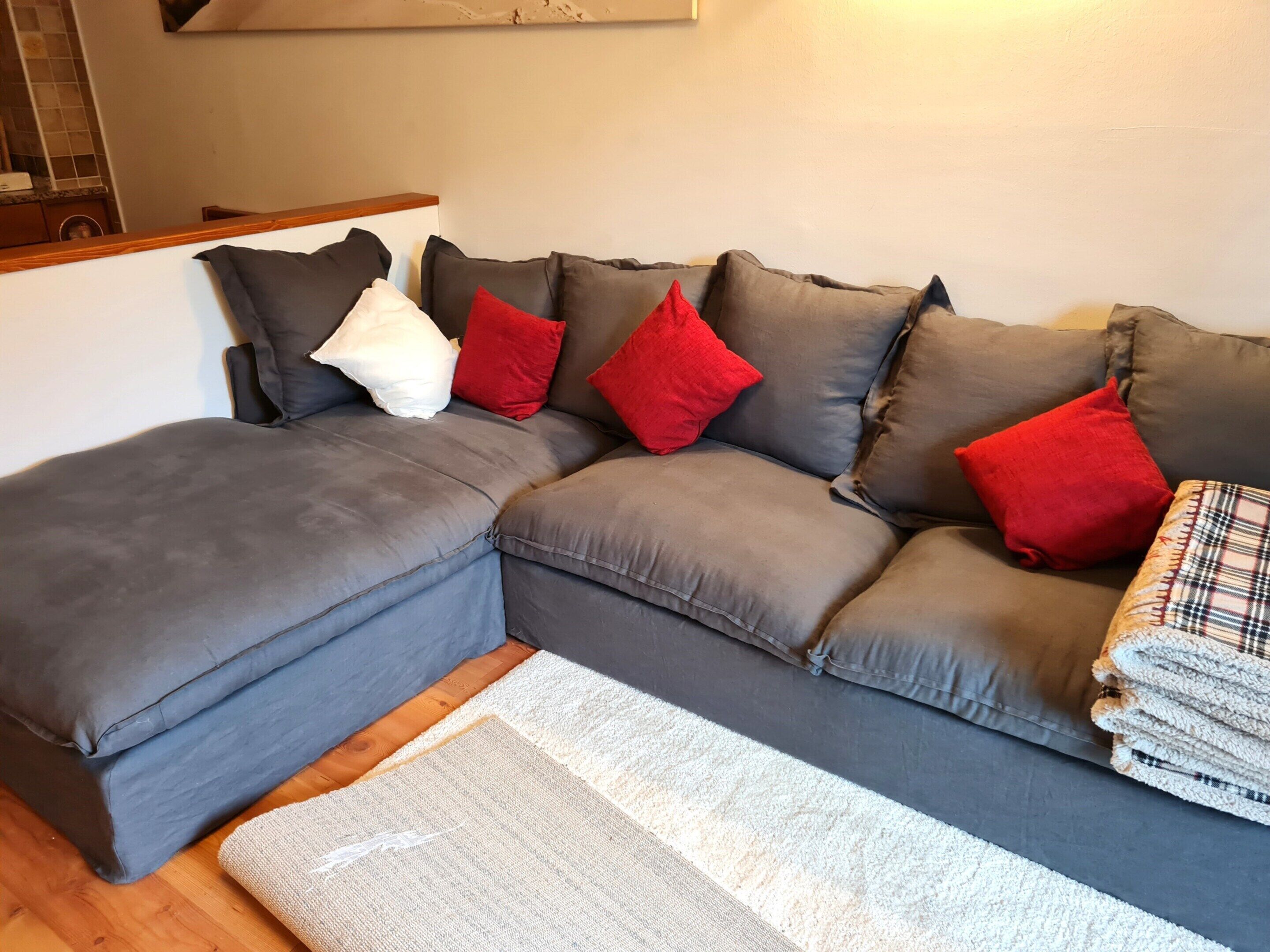 Um sofá modular com almofadas cinzas e almofadas decorativas vermelhas e brancas, um tapete branco e piso de madeira.