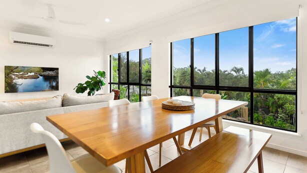 Un comedor moderno con una mesa y sillas de madera, ventanas amplias con vistas a zonas verdes, un sofá blanco con cojines y un cuadro en la pared.
