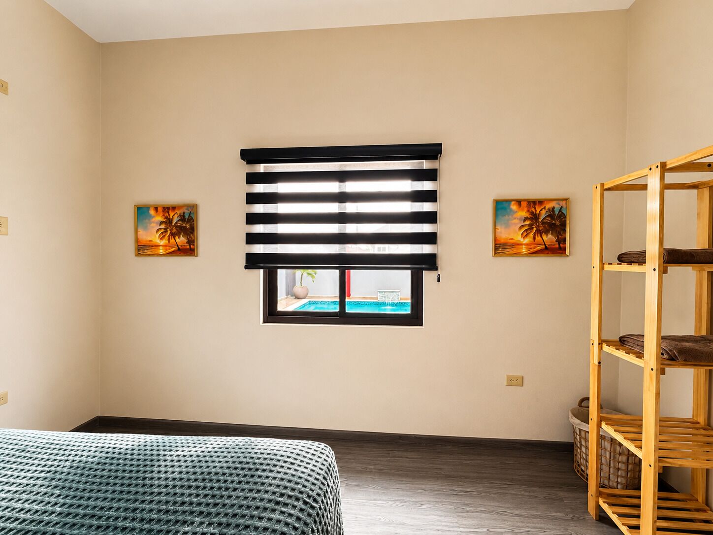 Een kamer met een bed, een raam met zonwering, een houten plank en twee schilderijen aan de muur.