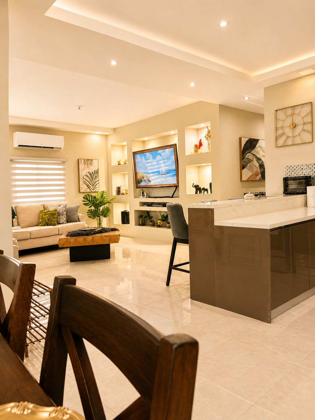 A modern kitchen with a central island, a dining area with wooden chairs, and a living room with a sofa and a TV.