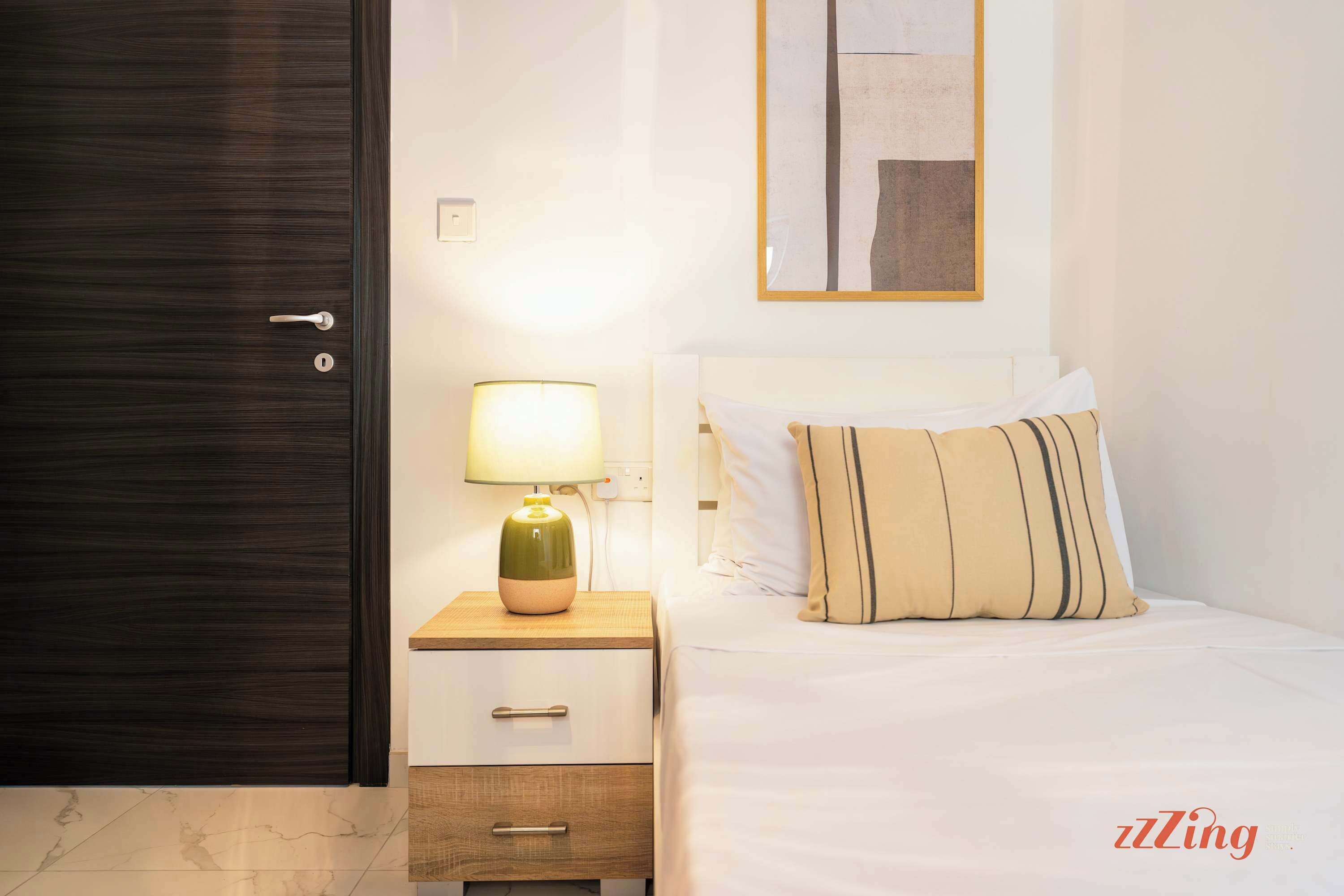 Habitación de hotel con cama, mesita de noche con lámpara, mesita con cajones, puerta de madera oscura y espejo adosado a la pared.