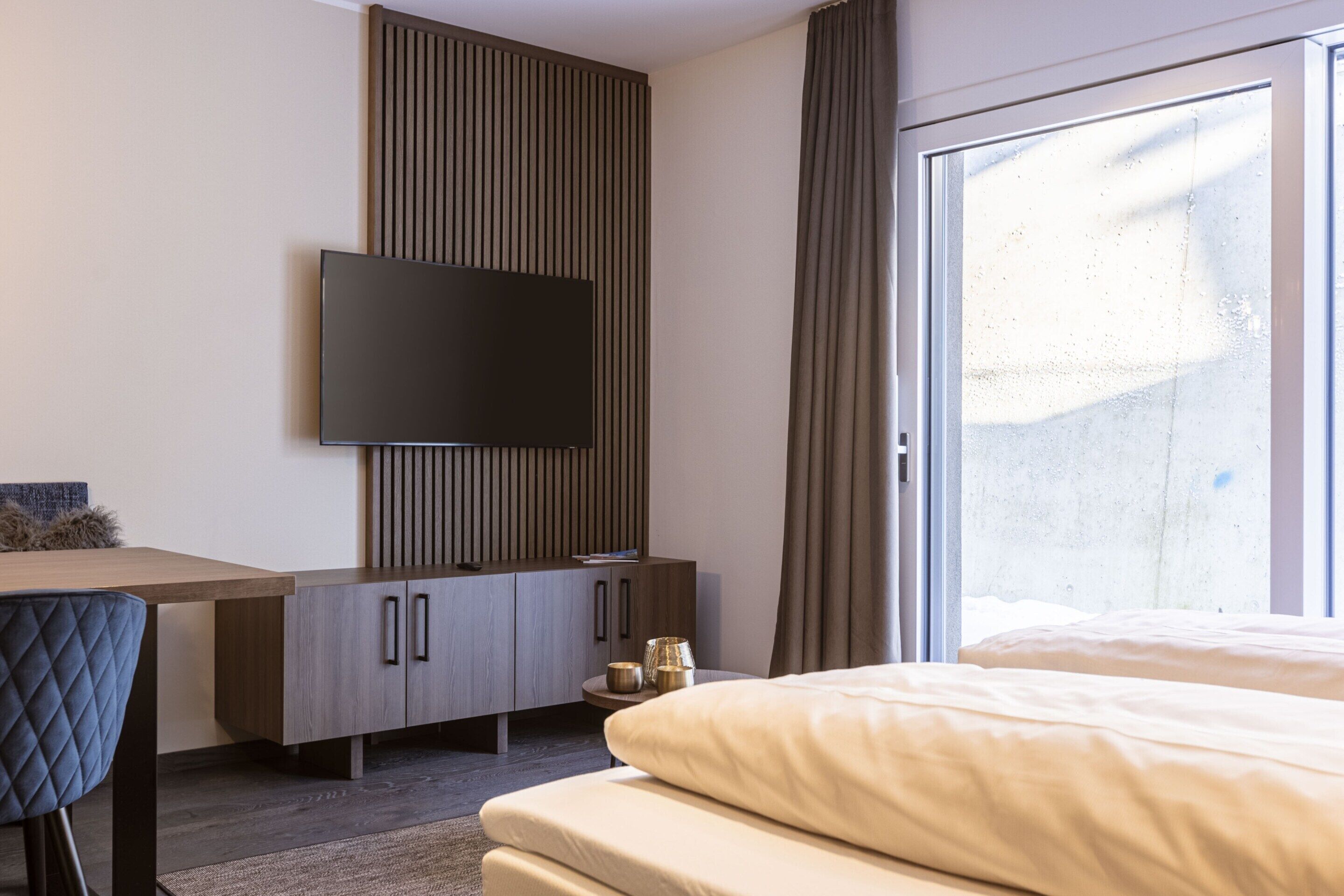 A modern hotel room with a flat-screen TV mounted on the wall, a wooden desk with a chair, a bed with white bedding, and a large window with curtains.