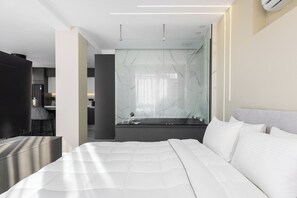 A modern hotel room with a large bed, a glass-enclosed bathroom, and a kitchenette area. - Ophelia Suites by halu! (Thessaloniki)