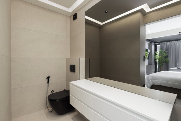 Un baño moderno con tocador blanco, un bidé negro y un espejo grande que refleja un dormitorio con cama y cortinas.