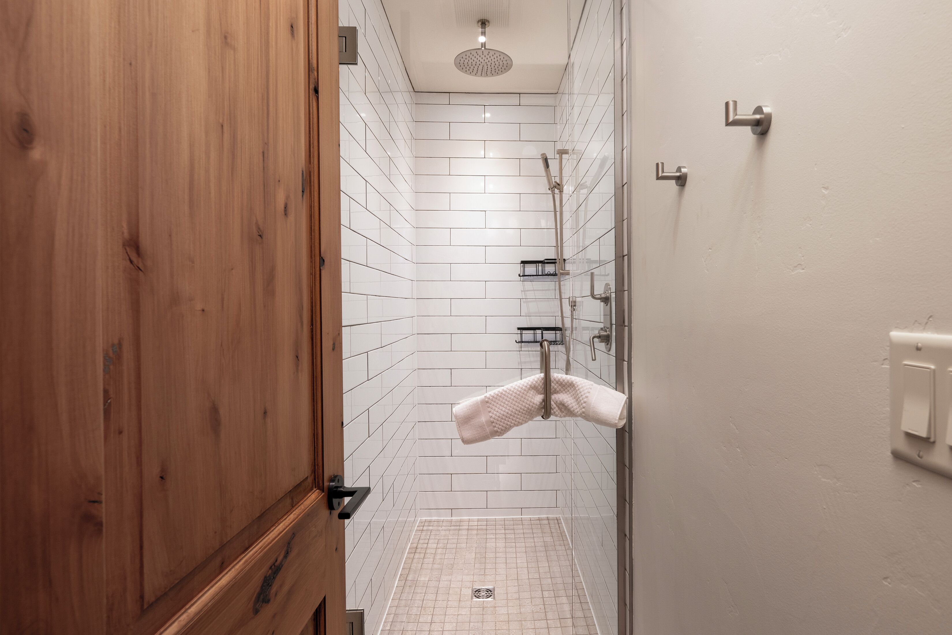 A shower area with white tiled walls, a wooden door, and a towel hanging on a rack.