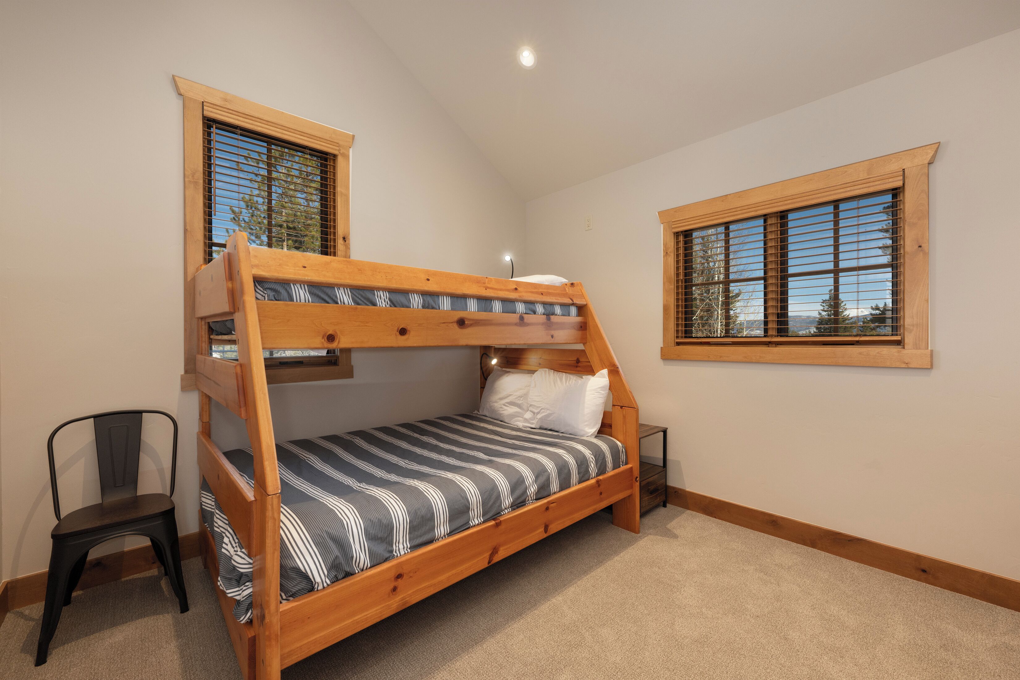 A bunk bed with a wooden frame, a striped bedspread, and a chair. There are two windows with wooden frames and blinds.