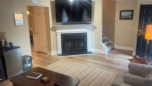 Ein Wohnzimmer mit Kamin, einem darüber montierten Fernseher, einem Couchtisch und einer Treppe, die in einen anderen Raum führt.