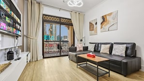 A modern living room with a black leather sofa, a coffee table, a TV mounted on the wall, and a balcony with a view of a cityscape. - Cozy 1BR Apartment | Close to Mall, Metro, and Dining (Dubai)