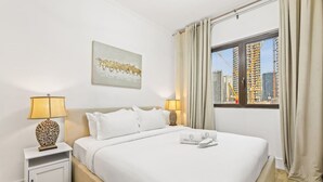 A hotel room with a bed, two bedside lamps, a window with curtains, a city view, and a painting on the wall. - Cozy 1BR Apartment | Close to Mall, Metro, and Dining (Dubai)