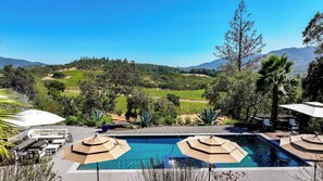 A pool area with lounge chairs and umbrellas, surrounded by a landscaped garden and a view of vineyards and hills.
