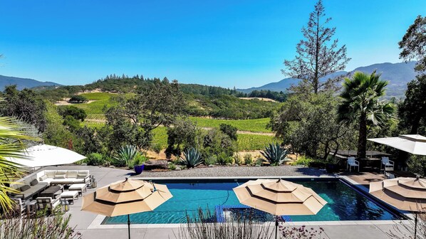 A pool area with lounge chairs and umbrellas, surrounded by a landscaped garden and a view of distant hills.