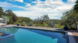 A clear swimming pool with a tiled edge, surrounded by a variety of plants and trees, and a wooden deck area.