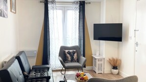 A modern living room with a sofa, armchair, and a coffee table with fruit. A flat-screen TV is mounted on the wall. - Norwich Delight Apartment (Romford)