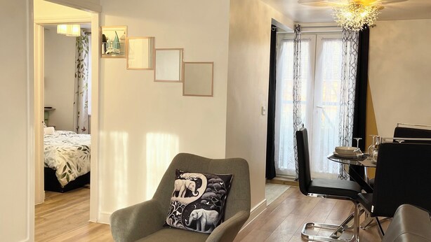 A modern living room with a grey armchair, a black sofa, a dining table with chairs, and a chandelier. There is a fruit basket on the table and a bed in the background. - Norwich Delight Apartment (Romford)