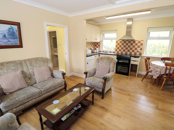 A living room with a sofa, armchairs, and a coffee table. There is a kitchen area with a stove, sink, and cabinets.