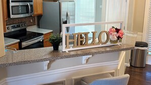 A kitchen with wooden cabinets, a microwave, a stove, and a refrigerator. There is a 'HELLO' sign and a vase with flowers on the counter.