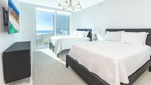 Ein Schlafzimmer mit einem großen Bett, einem kleineren Bett, einem Schreibtisch und Meerblick durch große Fenster.
