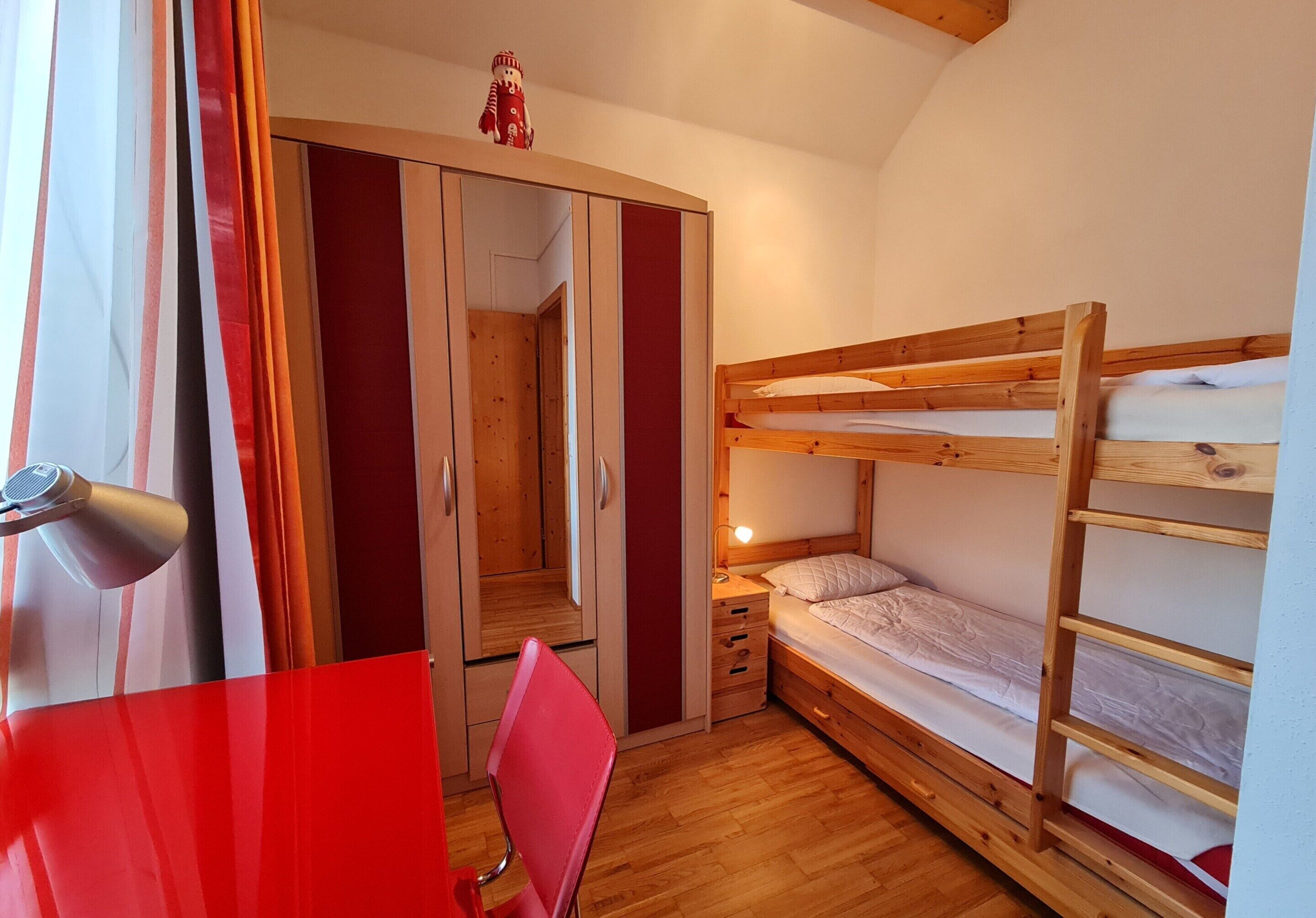 A bedroom with a bunk bed, a desk with a chair, a wardrobe, and a mirror.