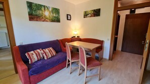 A cozy living room with a purple sofa, a wooden dining table, and two chairs. There are two framed pictures on the wall, one of a forest and another of a forest with a bench. - Beautiful Apartment in Bad Kleinkirchheim (Bad Kleinkirchheim)