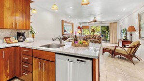 A modern kitchen with wooden cabinets, a dishwasher, and a dining area with a sofa and table. - Remodeled, A/C,-KS C2C - Sullivan Properties (Lahaina)