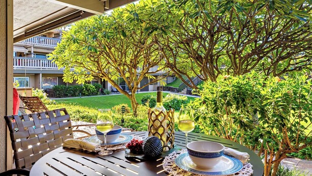 An outdoor dining area with a table set for two, surrounded by lush greenery and a view of a residential building. - Remodeled, A/C,-KS C2C - Sullivan Properties (Lahaina)