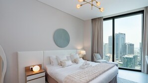 A modern hotel bedroom with a large bed, a round wall clock, a nightstand with a lamp, and a view of the city through large windows.