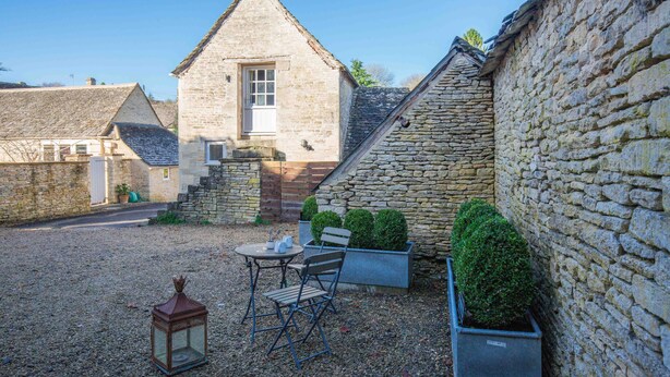 A courtyard with stone buildings, a gravel ground, a table with chairs, and potted plants. - Stunning contemporary cottage - The Bothy (Bibury)