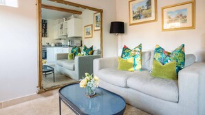 A living room with a sofa, coffee table, and a large mirror reflecting the kitchen area. - Stunning contemporary cottage - The Bothy (Bibury)
